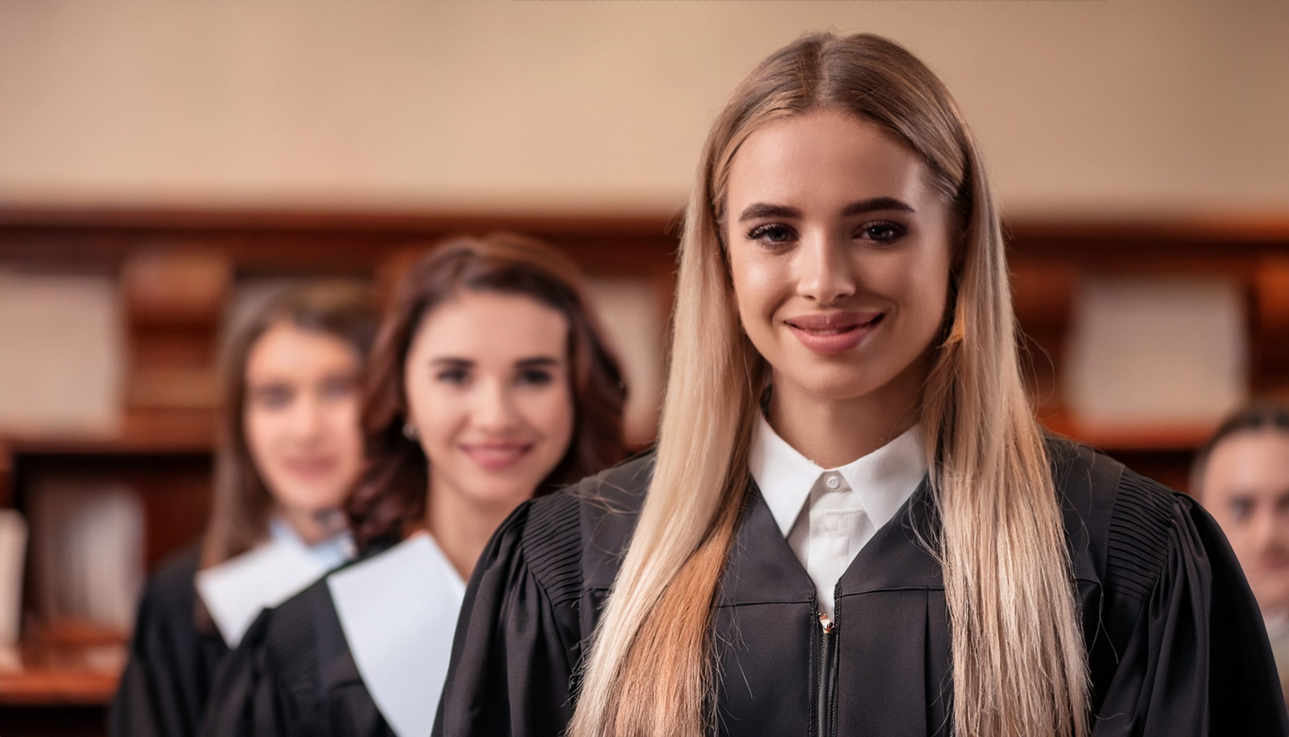 Portret uśmiechniętej młodej kobiety z długimi blond włosami, ubranej w czarną togę z białym żabotem. Za nią w rzędzie stoją dwie inne kobiety, również w togach, co wskazuje na ceremonię zakończenia studiów prawniczych lub grupę sędziów/adwokatów.
