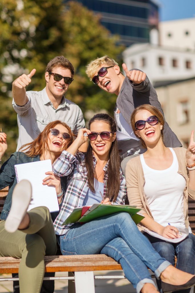 Grupa pięciorga uśmiechniętych studentów siedzi na ławce na zewnątrz budynku. Wszyscy mają na sobie okulary przeciwsłoneczne, trzymają notatniki i foldery. Dwóch mężczyzn w tle pokazuje gest kciuka w górę.
